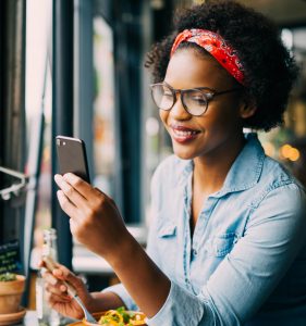 Young African woman smiling and reading texts on a cellphone while sitting alone at a counter in a café enjoying a meal.